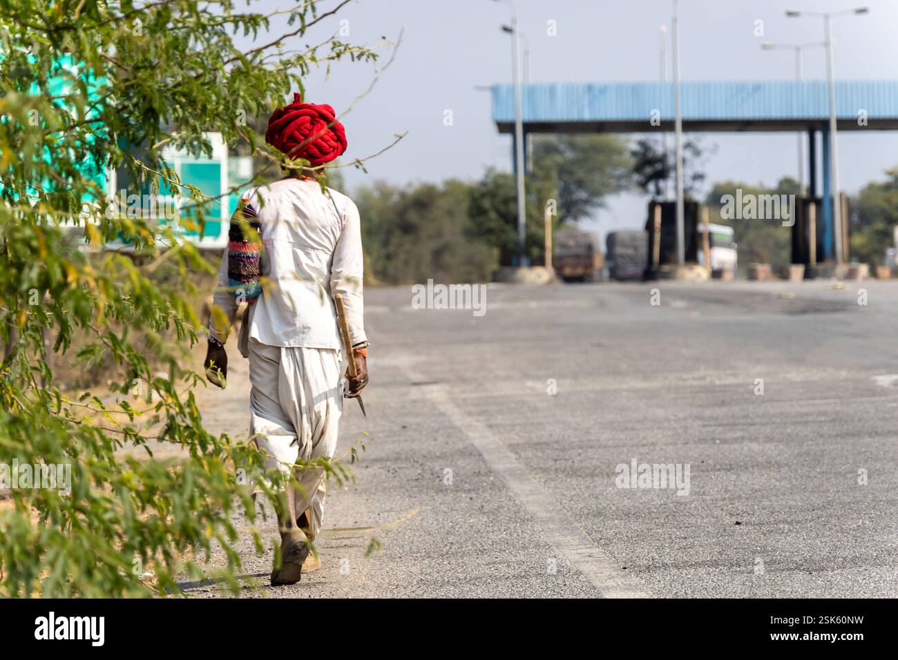 hirte in traditioneller Kleidung mit Turban und Stock und Spaziergang auf der Asphaltstraße in der Nähe des Dorfes am Tag Stockfoto