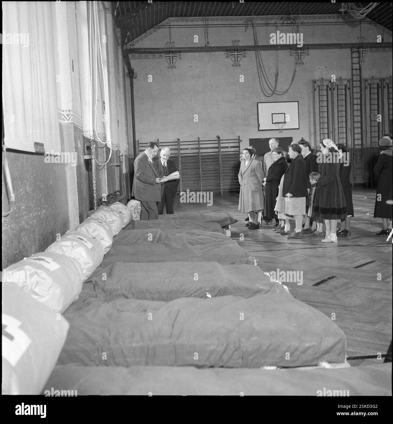 Gemeindepräsident Zwissig, Verteilung der neun Betten; Zwischenlager im Turnsaal#Gemeindepräsident Zwissig, Verteilung von neun Betten; Zwischenlager im Turnsaal--- SRK-Betten-Aktion in Siders 1954#Schweizerisches Rotes Kreuz 'Betten' Hilfsaktion in Sierre 1954- RDB VON DUKAS Stockfoto