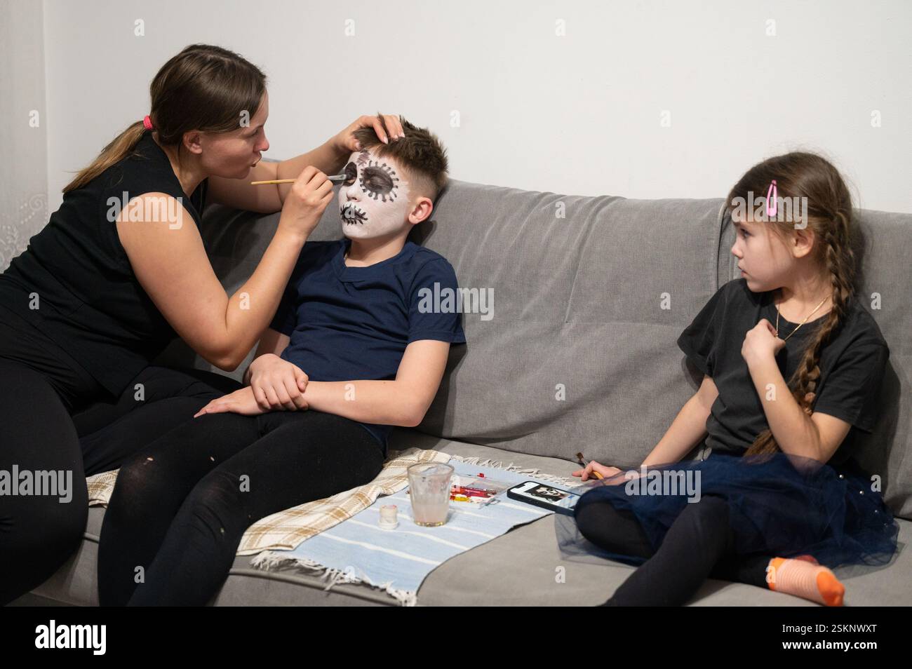 Eine Frau trägt sorgfältig Gesichtsfarbe auf ihren Sohn auf, um ein Schädeldesign zu kreieren. Ihre Tochter beobachtet von der Couch aus. Eine Szene der Kreativität der Familie und Hal Stockfoto