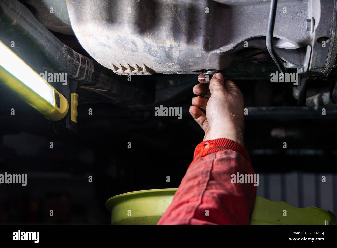Nahaufnahme eines Mechanikers, der die Ölablassschraube aus der Ölwanne eines Autos herausdreht, um altes Motoröl während eines Ölwechsels abzulassen. Der Arbeiter trägt einen Stockfoto