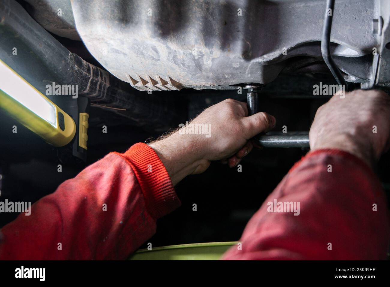 Nahaufnahme der Hände eines Mechanikers mit einem Schraubenschlüssel zum Lösen der Ölablassschraube an der Ölwanne eines Autos während eines Motorölwechsels. Der Arbeiter, gekleidet in einer Re Stockfoto
