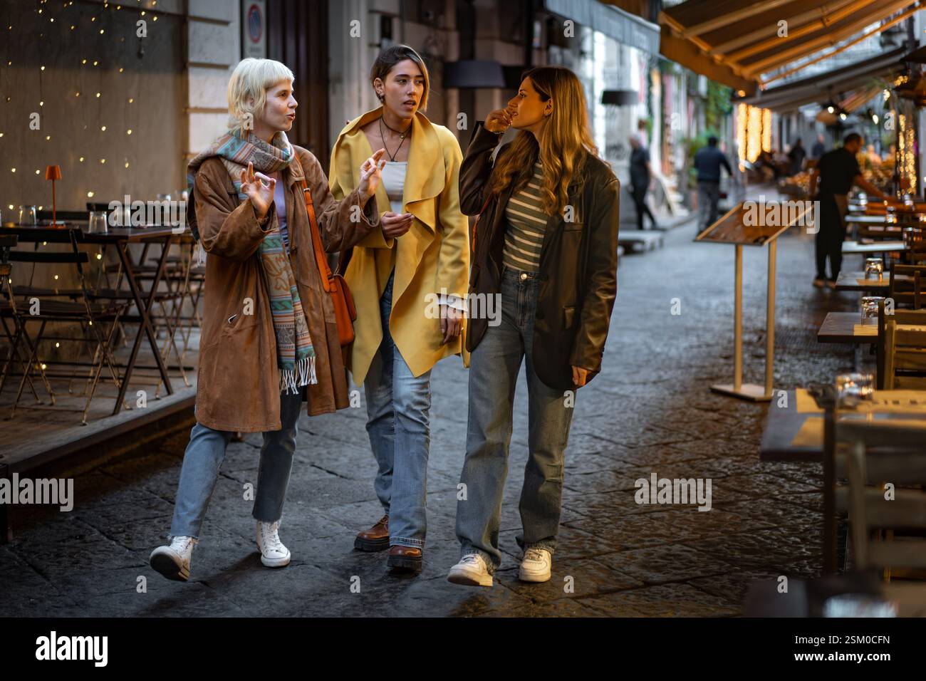 Drei modische junge Frauen schlendern und plaudern entlang einer charmanten Straße, beleuchtet von warmen Abendlichtern. Die Szene fängt eine entspannte an ein Stockfoto