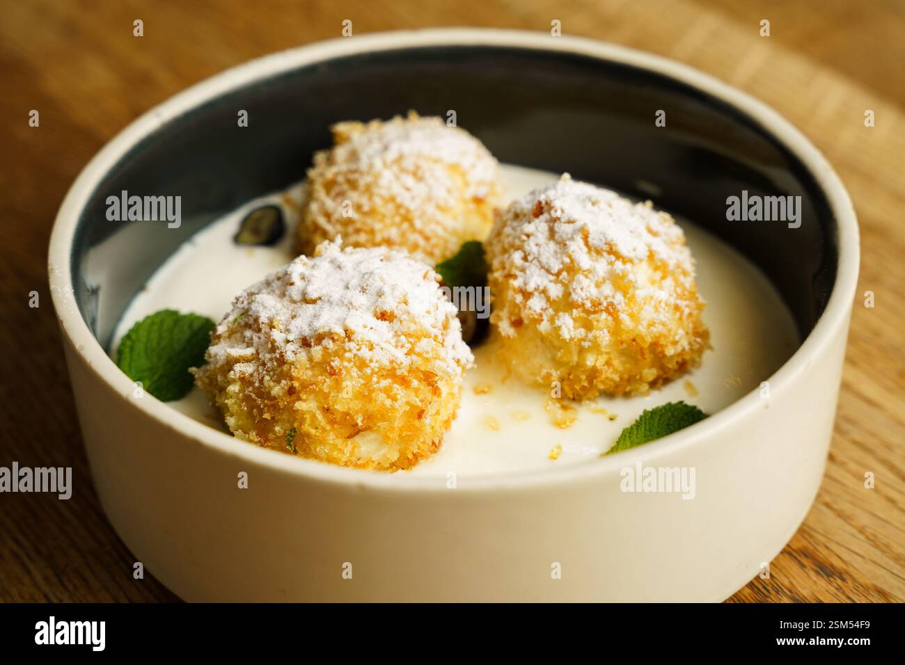 Goldene, knusprig gebratene Hüttenkäse-Knödel, mit Puderzucker bestäubt, serviert in einer Schüssel mit cremiger Vanillesauce. Garniert mit frischen Minzblättern Stockfoto