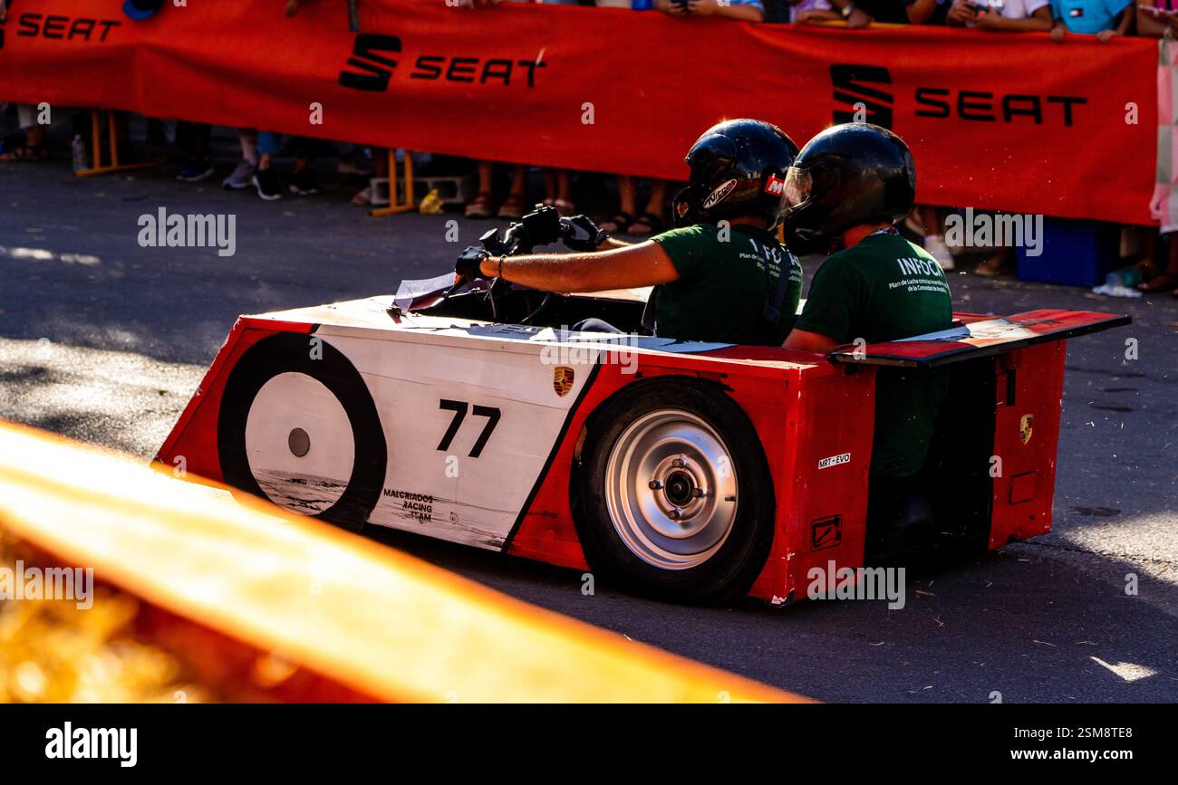 Aufregende Soapbox-Rennszene mit dem Custom Red and White Car Nr. 77 und zwei Helmfahrern, die sich in der Masse und in Sponsoren befinden Stockfoto