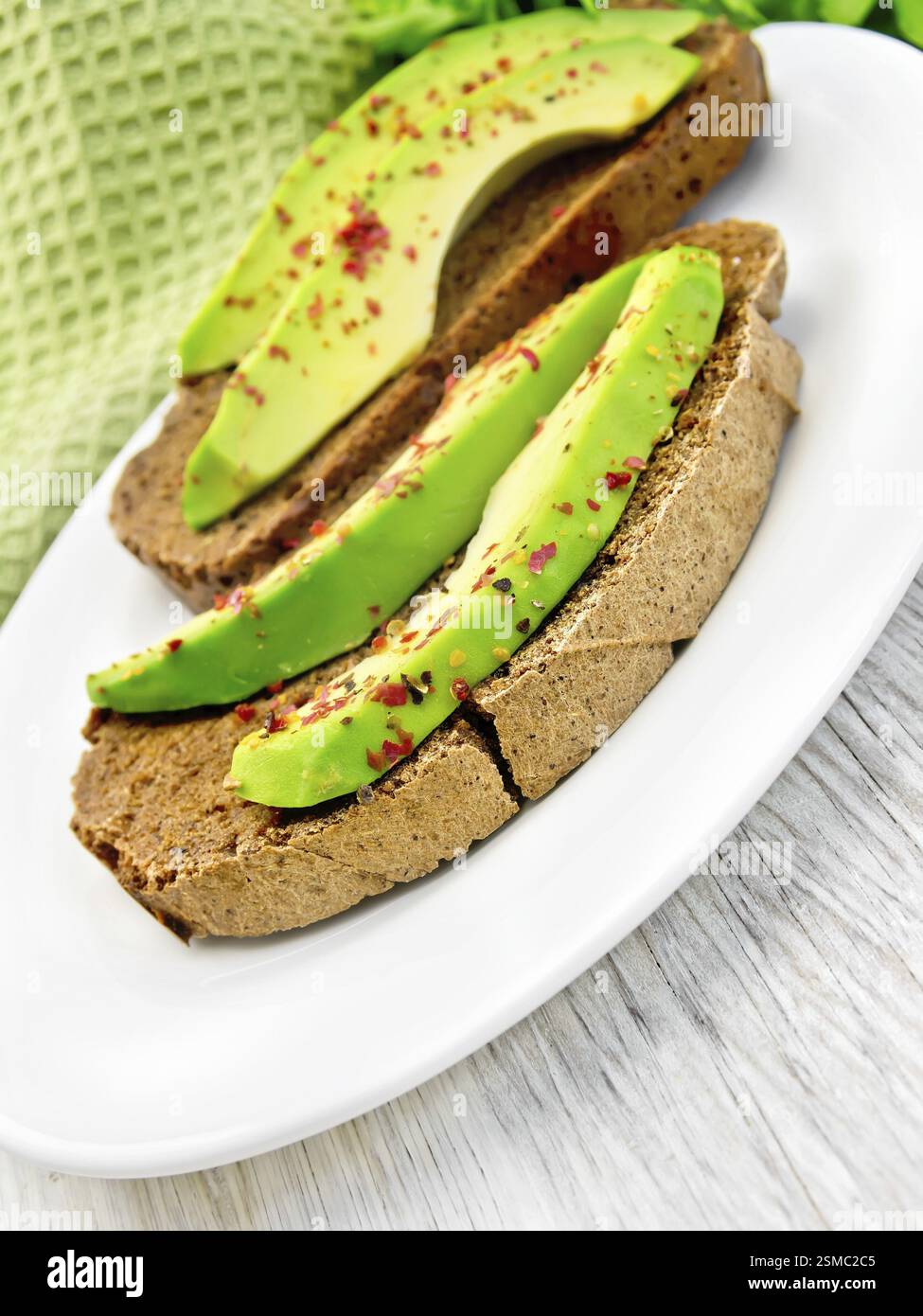 Zwei Scheiben Roggenbrot mit Scheiben Avocado und Pfeffer auf einem Teller, Servietten auf dem Hintergrund Licht Holzbretter Stockfoto