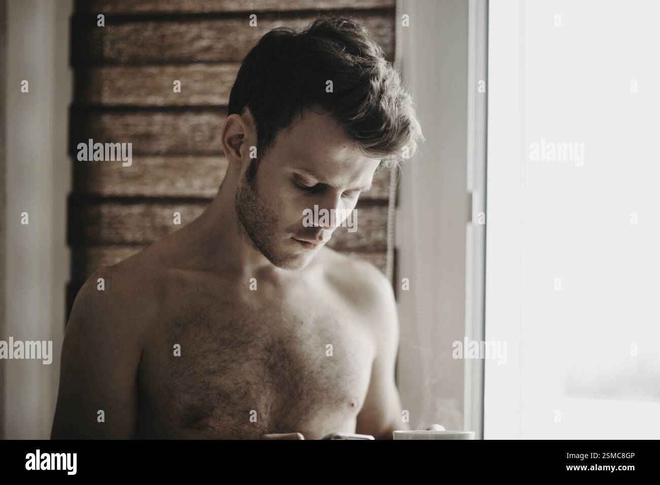 Ein Mann, der eine Kaffeetasse hält, in sanftem Licht an einem Fenster steht und besinnt Stockfoto