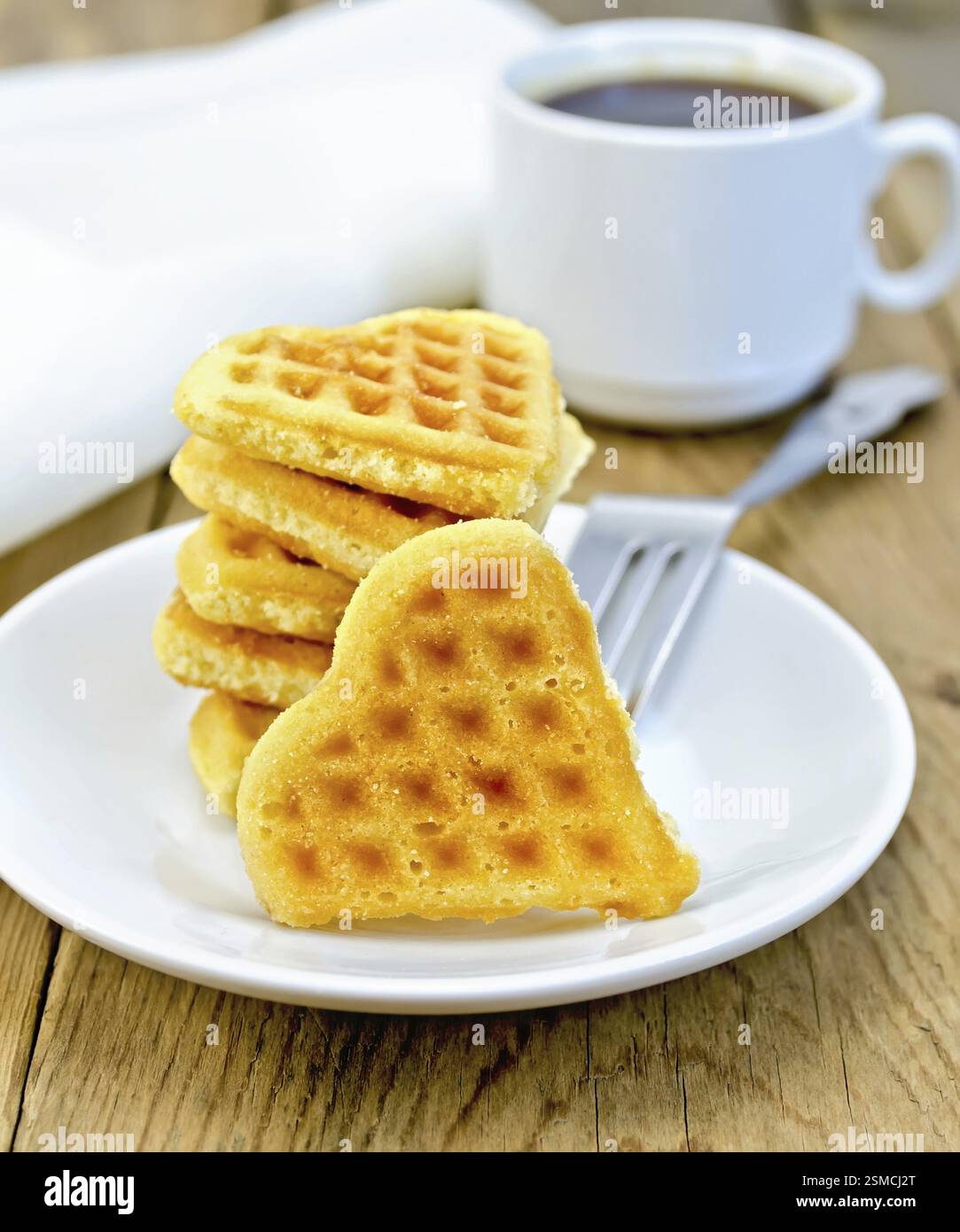 Ein Stapel von Cookies in der Form eines Herzens mit einer Gabel auf einem weißen Teller, Tasse und Serviette auf einem Hintergrund von Holzbrettern Stockfoto