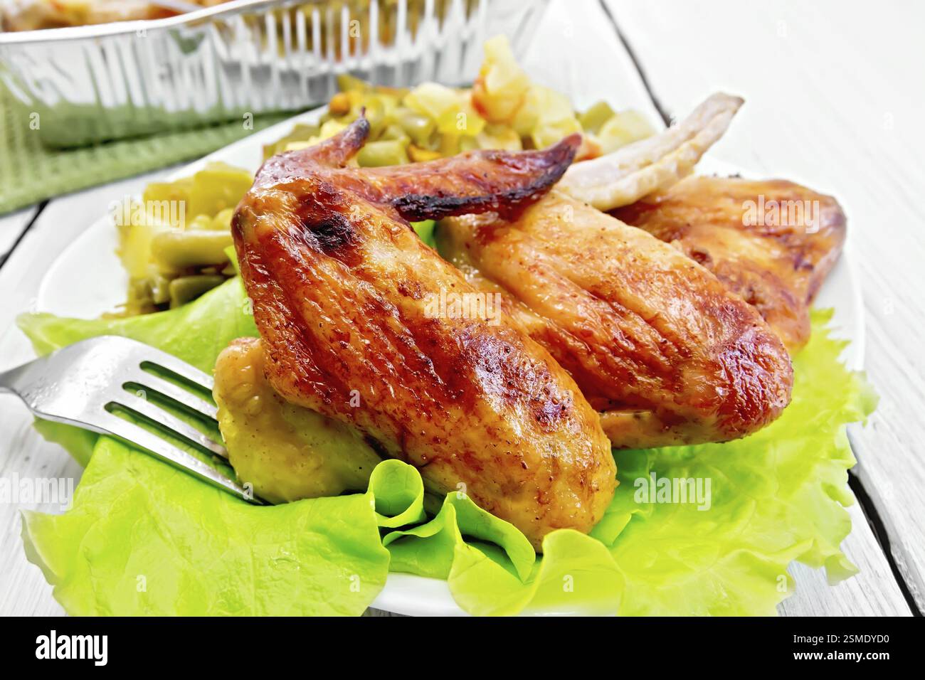 Gebratene Hähnchenflügel mit Gemüse und Salatblättern auf einem Teller, Gabel auf dem Hintergrund helle Holzbretter Stockfoto