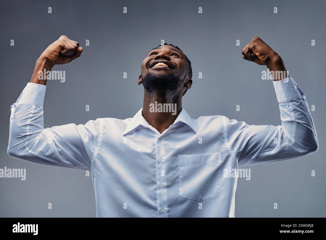 Studio-Porträt eines jungen afrikanischen Geschäftsmannes, der seinen Bizeps beugt und mit einem großen Lächeln aufblickt und in seinem weißen Hemd Selbstvertrauen und Kraft ausdrückt Stockfoto