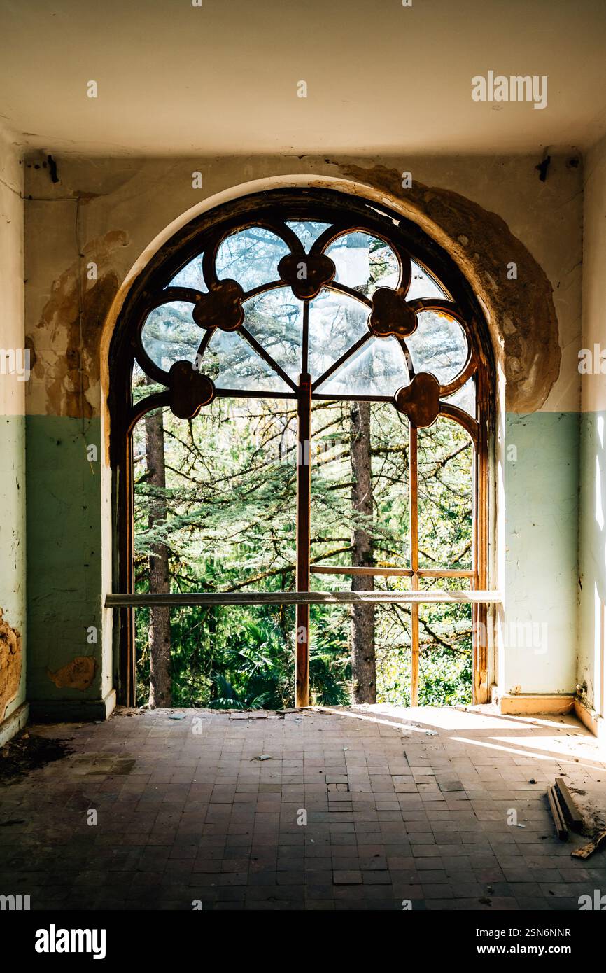Verzierte Vintage-Fenster in einem verlassenen Herrenhaus mit abblätternder Farbe und bewachsenen Bäumen, die draußen sichtbar sind, erinnern an Nostalgie und historischen Verfall. Stockfoto