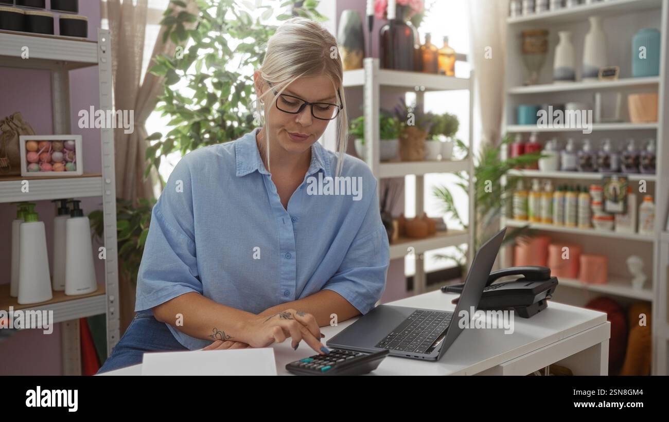 Blonde Frau mit Brille arbeitet an einem Laptop und Rechner in einem Wohnkultur-Geschäft mit Regalen gefüllt mit verschiedenen Gegenständen und Pflanzen im Hintergrund Stockfoto