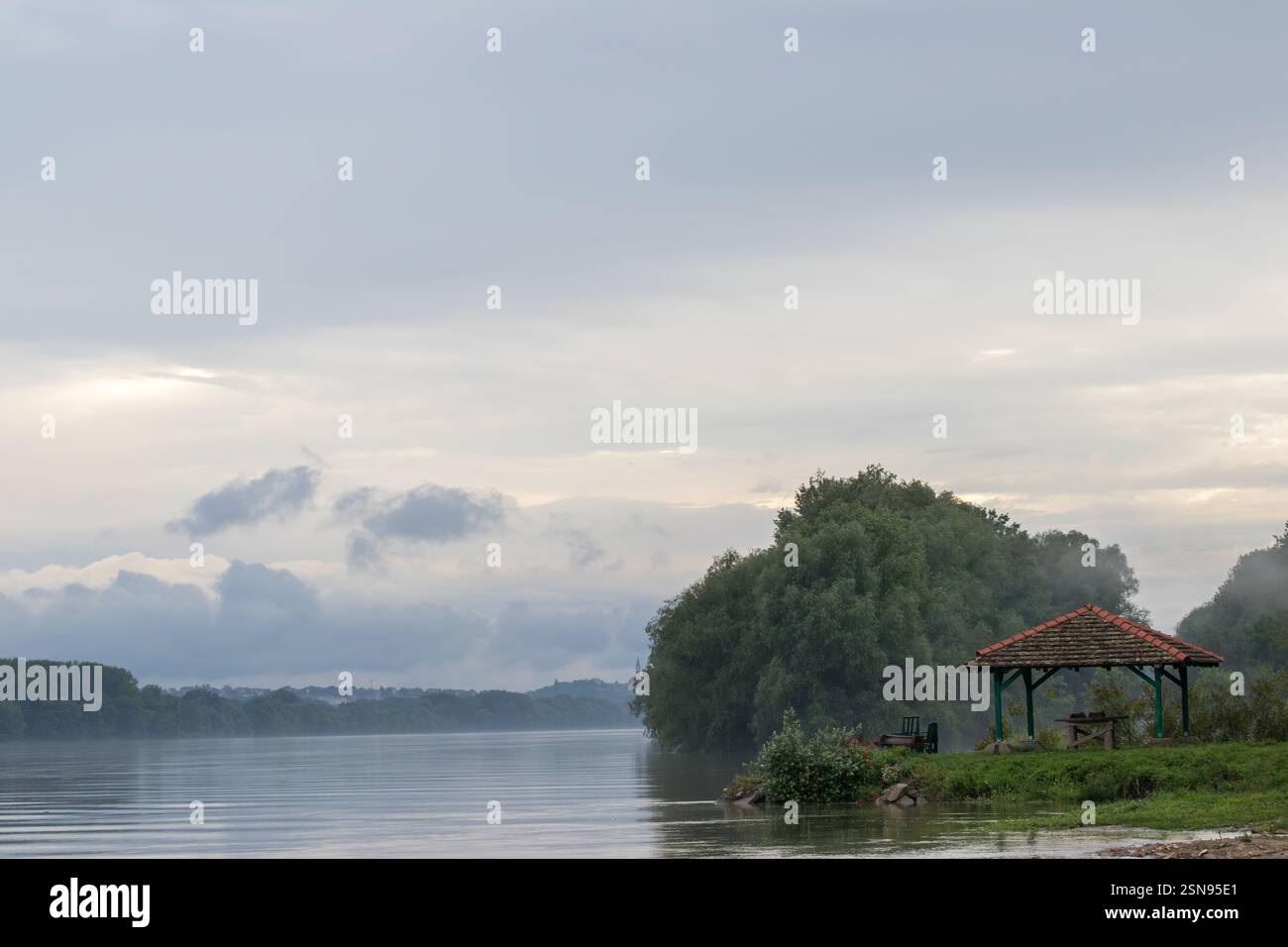 Wunderschöner Blick auf den Fluss mit einem hölzernen Pavillon, üppigen grünen Bäumen und bewölktem Himmel. Stockfoto