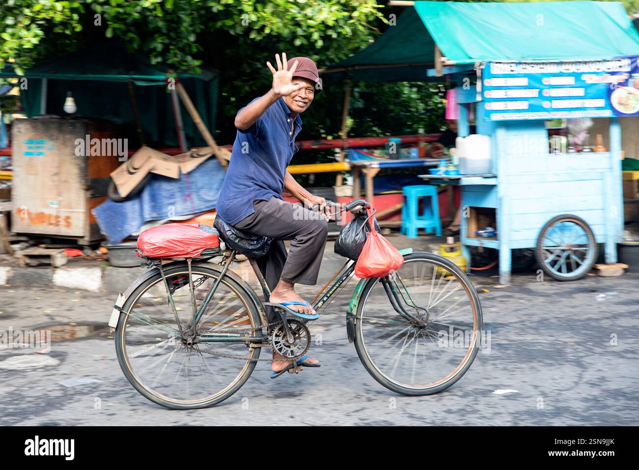 Ein lächelnder Mann winkt mit der Hand auf dem Fahrrad und trägt Plastiktaschen in Chinatown, Jakarta, Java, Indonesien Stockfoto