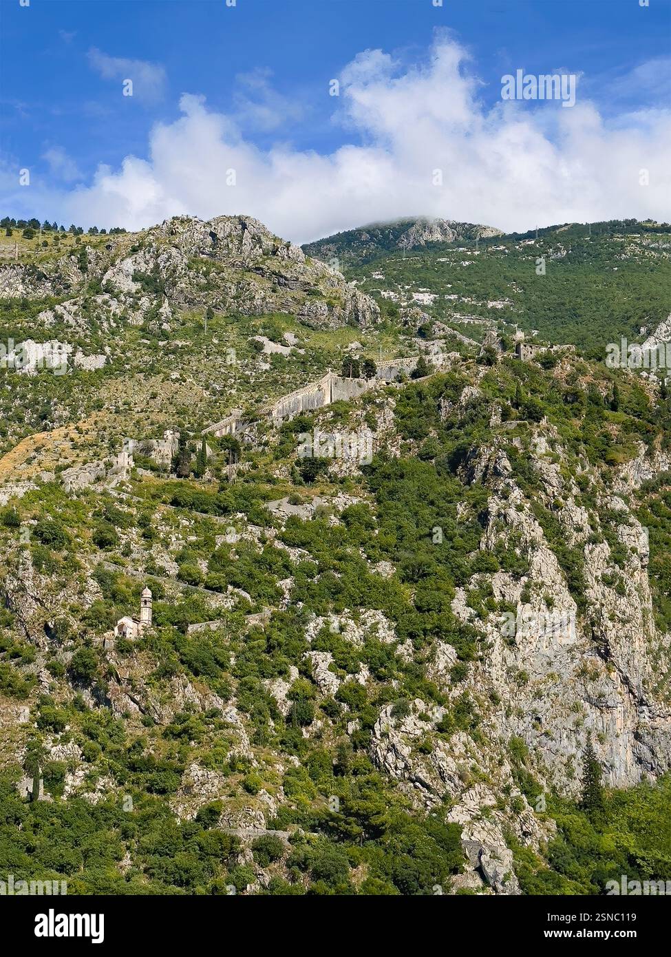 Kotor, Montenegro - 2. Juli 2024: Kirche unserer Lieben Frau von Remedy auf dem grünen bewaldeten Berg und darüber die Festungswälle unter blauer Wolkenlandschaft Stockfoto
