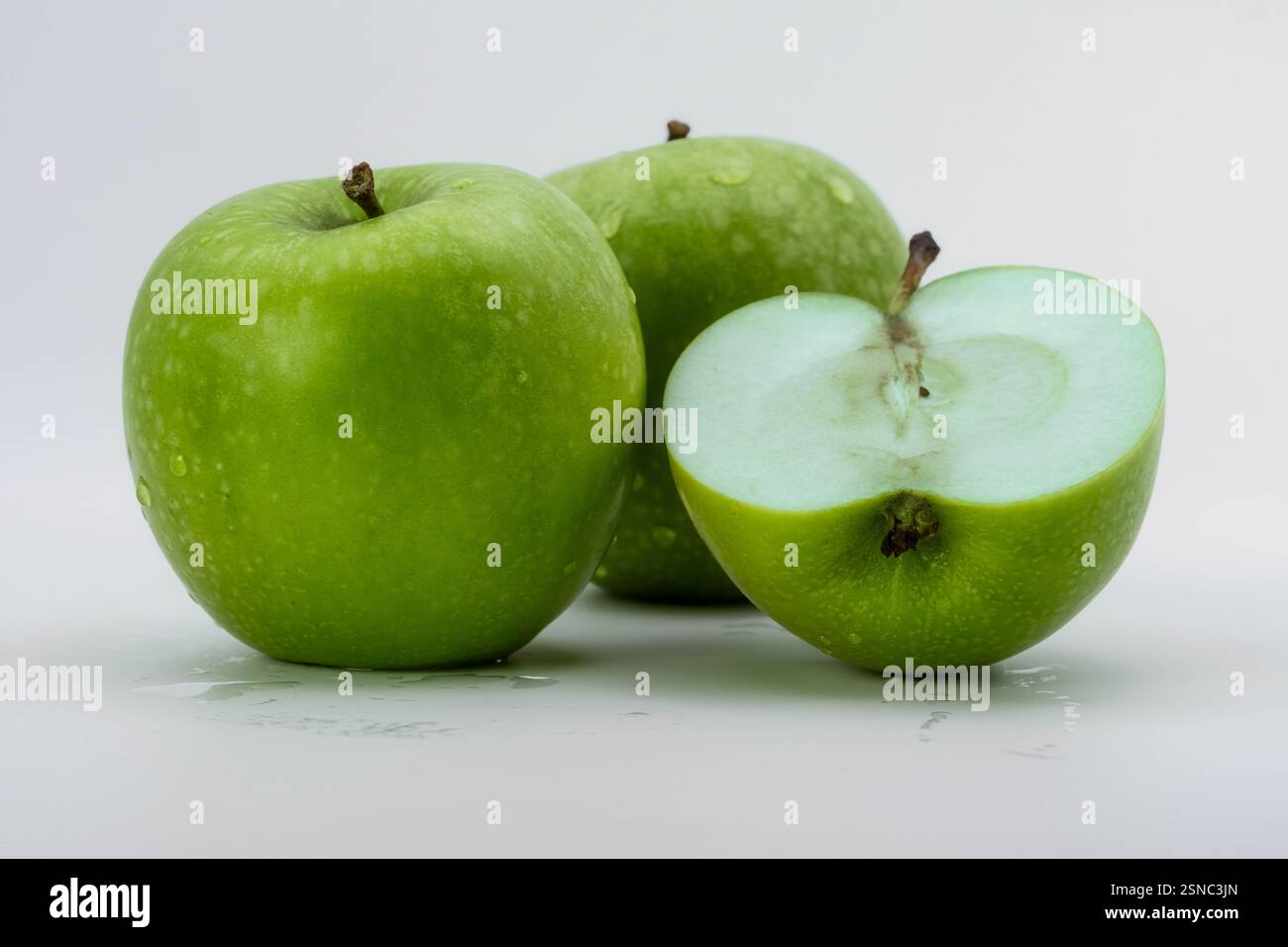Zwei ganze grüne Äpfel mit einem halbgeschnittenen Stück auf weißem Hintergrund. Eine knusprige und frische Komposition, perfekt für Gesundheit, Ernährung und Lebensmittelkonzepte. Stockfoto