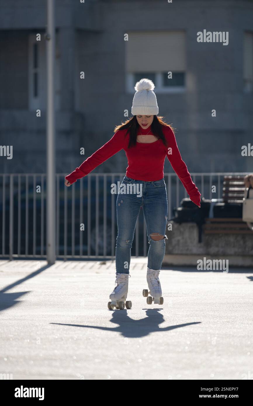 Eine junge Frau mit einer weißen Mütze, einem roten Rollkragenpullover und einer zerrissenen Jeans genießt das Rollschuhlaufen in einem Stadtpark und zeigt einen trendigen und aktiven urba Stockfoto