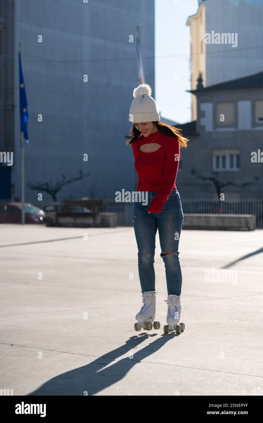 Die junge Frau mit rotem Pullover und weißer Mütze macht an einem sonnigen Tag in einer städtischen Umgebung Rollschuhlaufen und zeigt ein unbeschwertes und aktives Leben Stockfoto