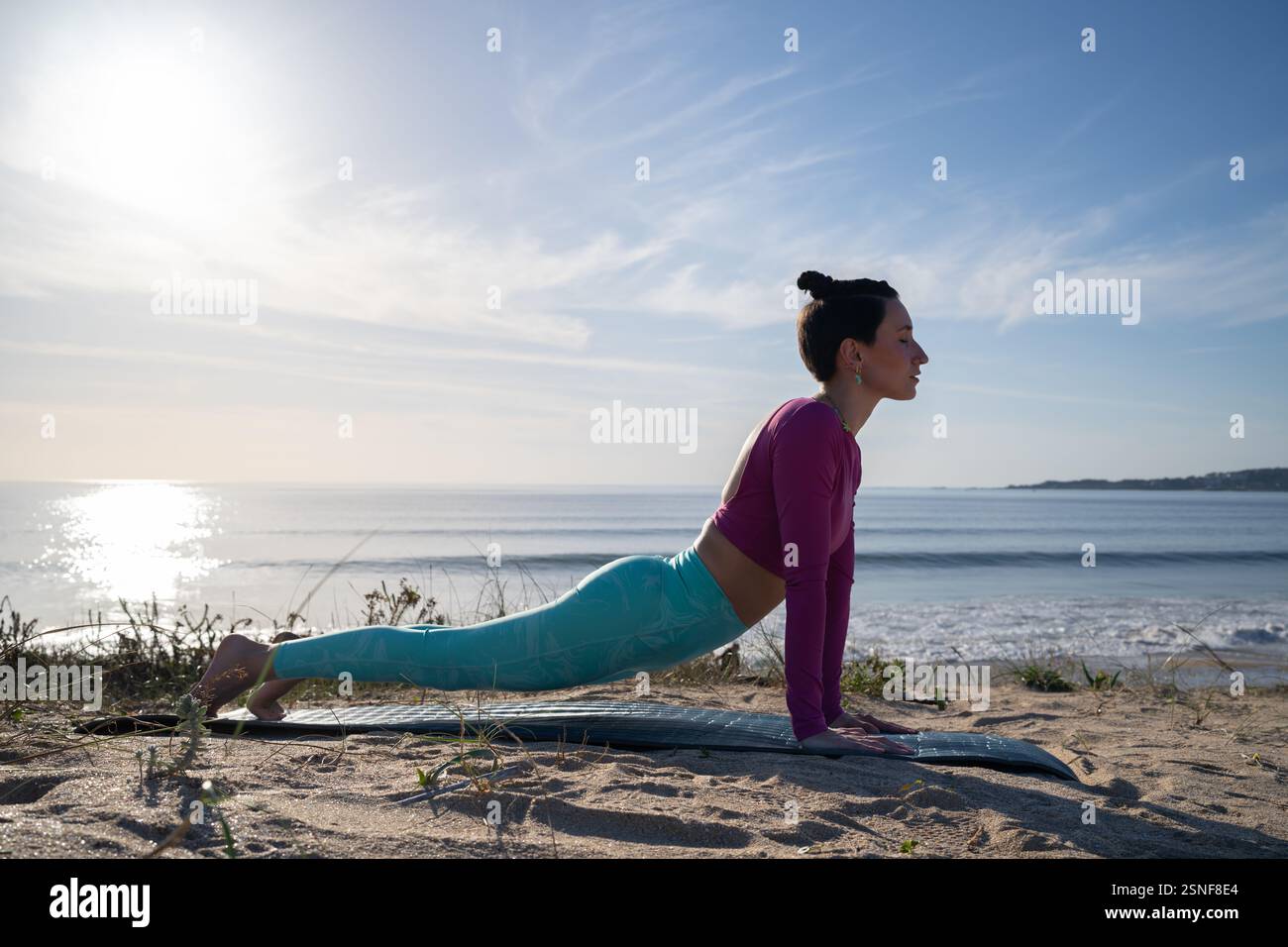 Junge Frau in Sportbekleidung übt Yoga auf einer Matte am Strand an einem sonnigen Tag, die nach oben gerichtete Hundestellung, fördert Gesundheit und Wellness Stockfoto