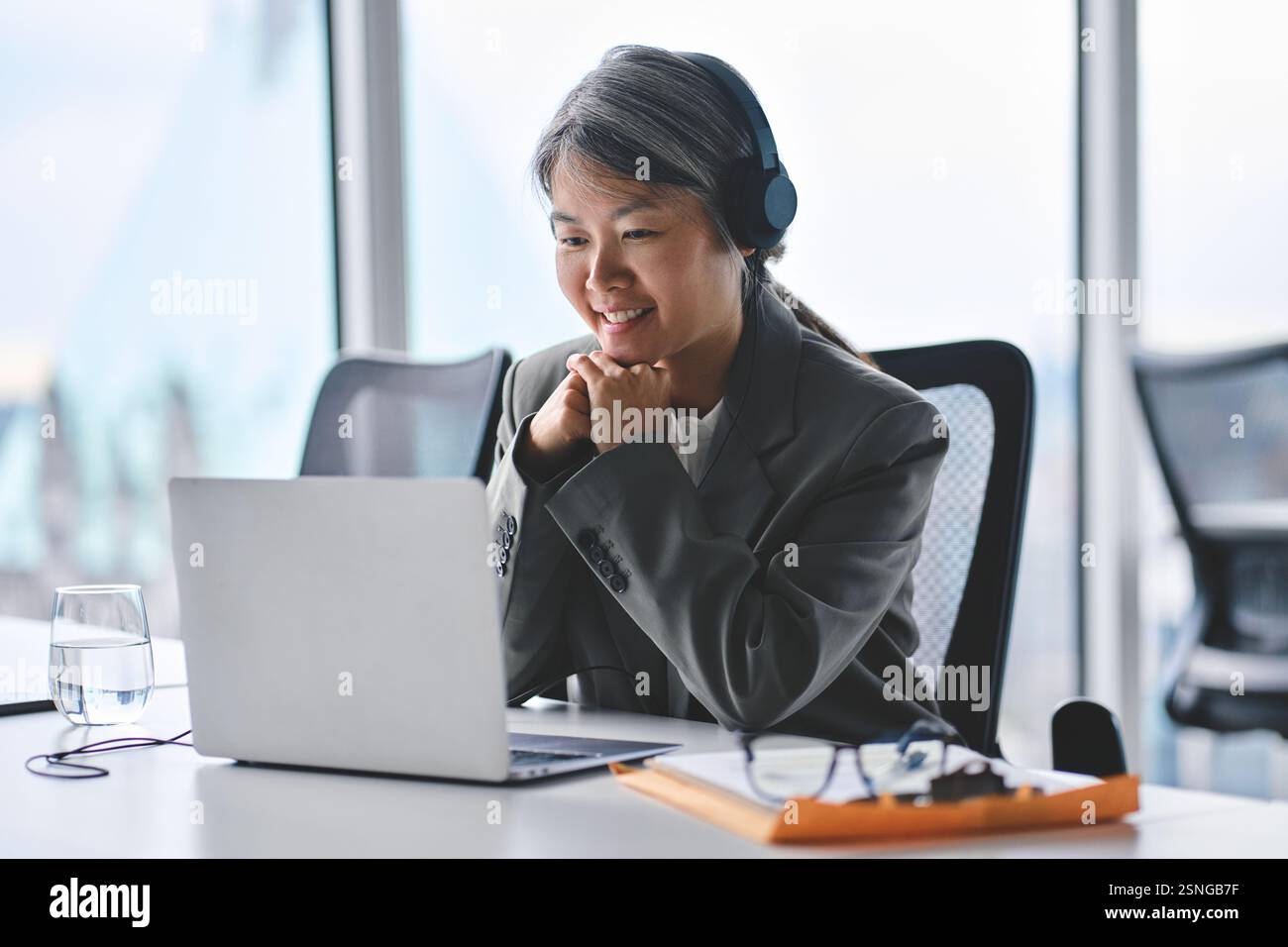 Beschäftigte, glückliche, reife asiatische Geschäftsfrau in Kopfhörern mit Laptop. Stockfoto