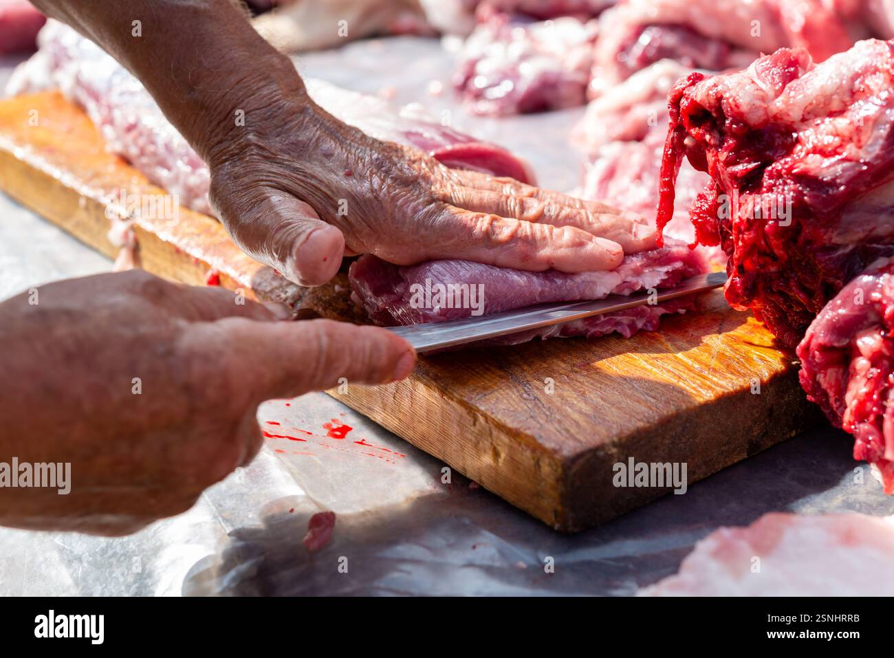 Die Hände eines erwachsenen Mannes schneiden rohes Schweinefleisch mit einem Messer auf einem hölzernen Schneidebrett draußen. Stockfoto