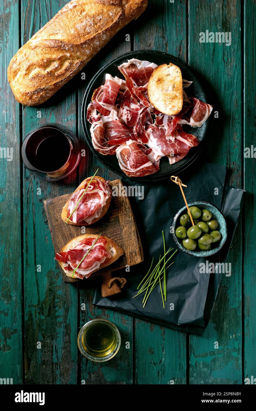 In Scheiben geschnittener spanischer Schinken Jamon Iberico paleta auf schwarzem Teller. Schüssel grüne Oliven, frisches und geröstetes Weißbrot, Olivenöl, Glas Rotwein, Tapas. Alt Stockfoto