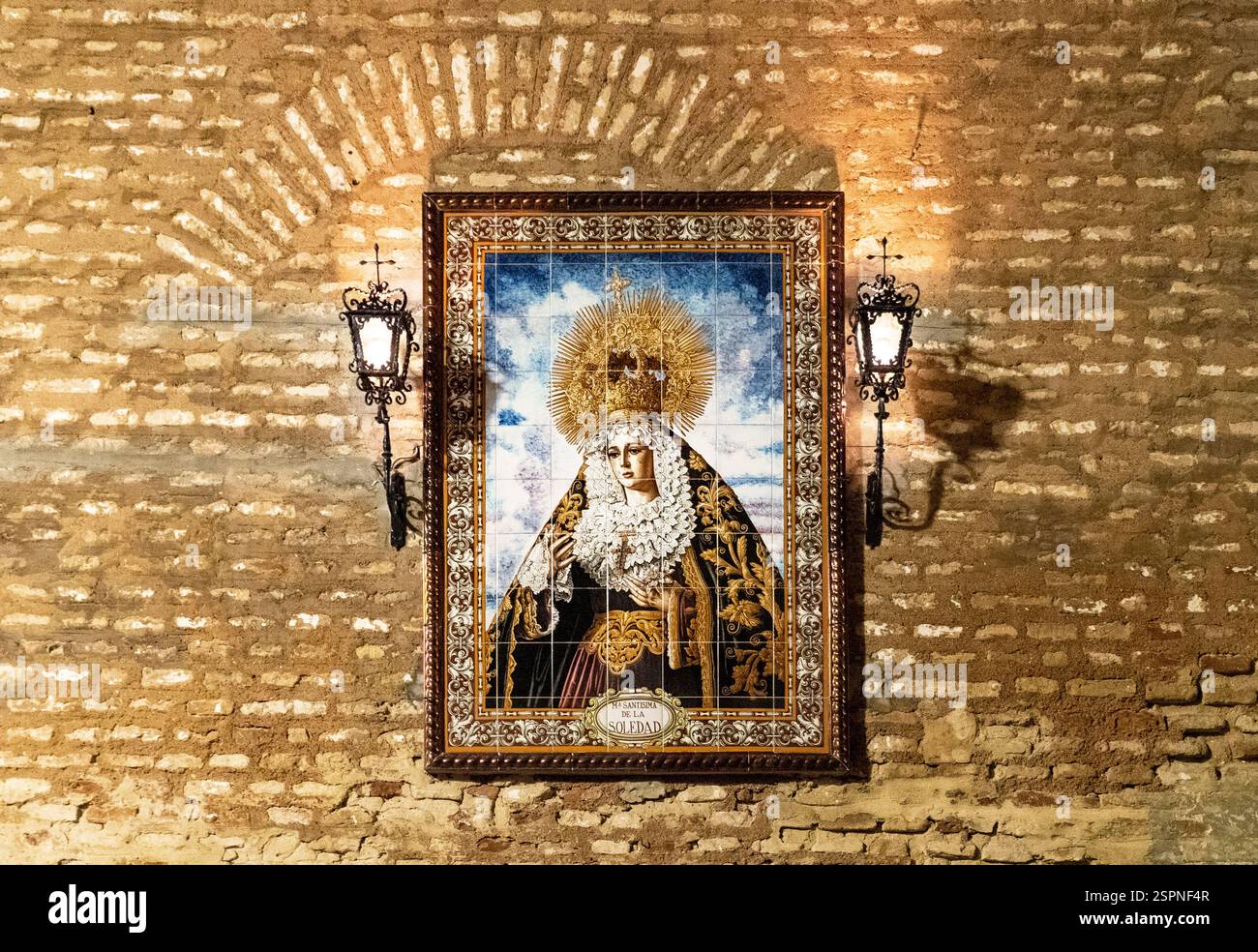 Ein religiöses Kunstwerk, das eine Figur mit Krone und aufwendiger Kleidung darstellt, gerahmt und auf einer strukturierten Ziegelwand montiert. Zwei kunstvolle Wandlampen leuchten Stockfoto