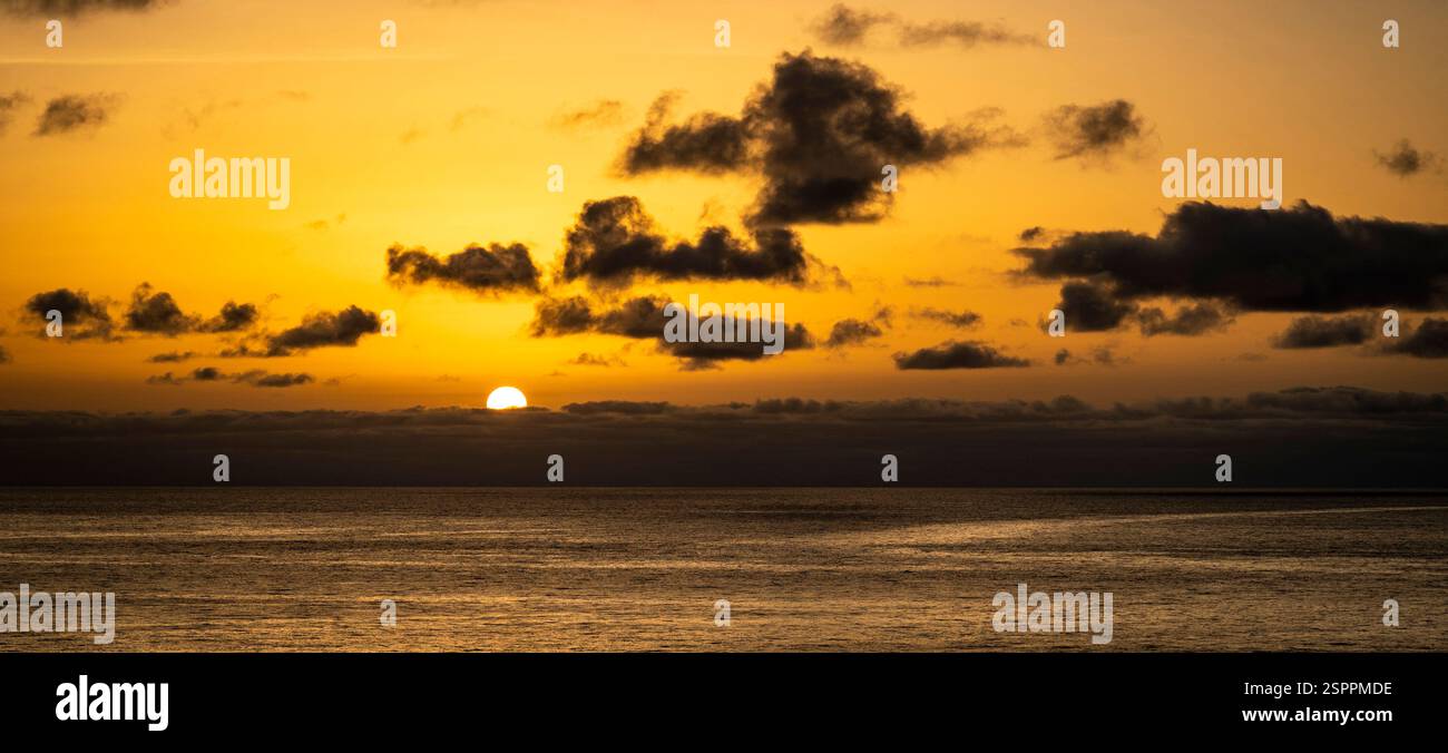 Ein ruhiger Sonnenuntergang über dem Meer, mit der Sonne teilweise hinter Wolken versteckt. Der Himmel ist in Orange- und Gelbtönen gemalt und reflektiert die Ruhe Stockfoto