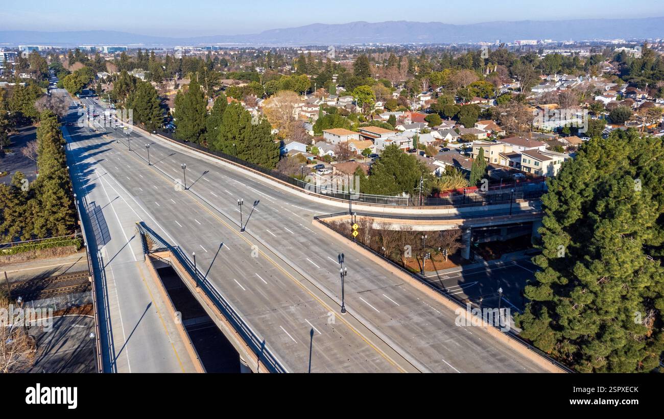 Aus der Vogelperspektive auf eine leere Überführung in einem Vorstadtviertel, umgeben von Bäumen und Wohnhäusern, mit fernen Bergen unter einem klaren blauen Himmel Stockfoto