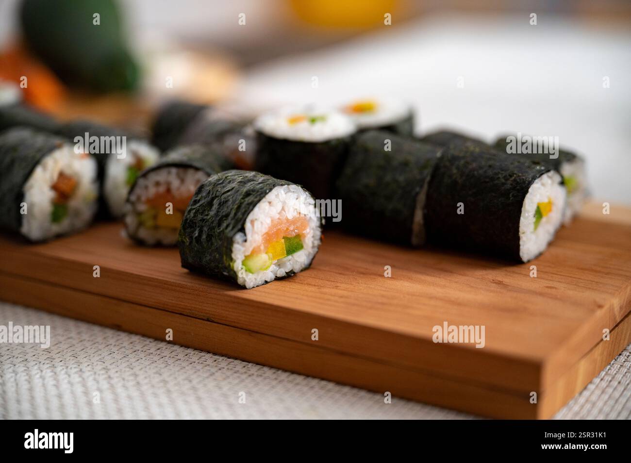 Eine Nahaufnahme frisch zubereiteter Sushi-Brötchen mit Lachs, Gurke und Paprika, in Nori gewickelt und auf einem Holzbrett platziert. Stockfoto