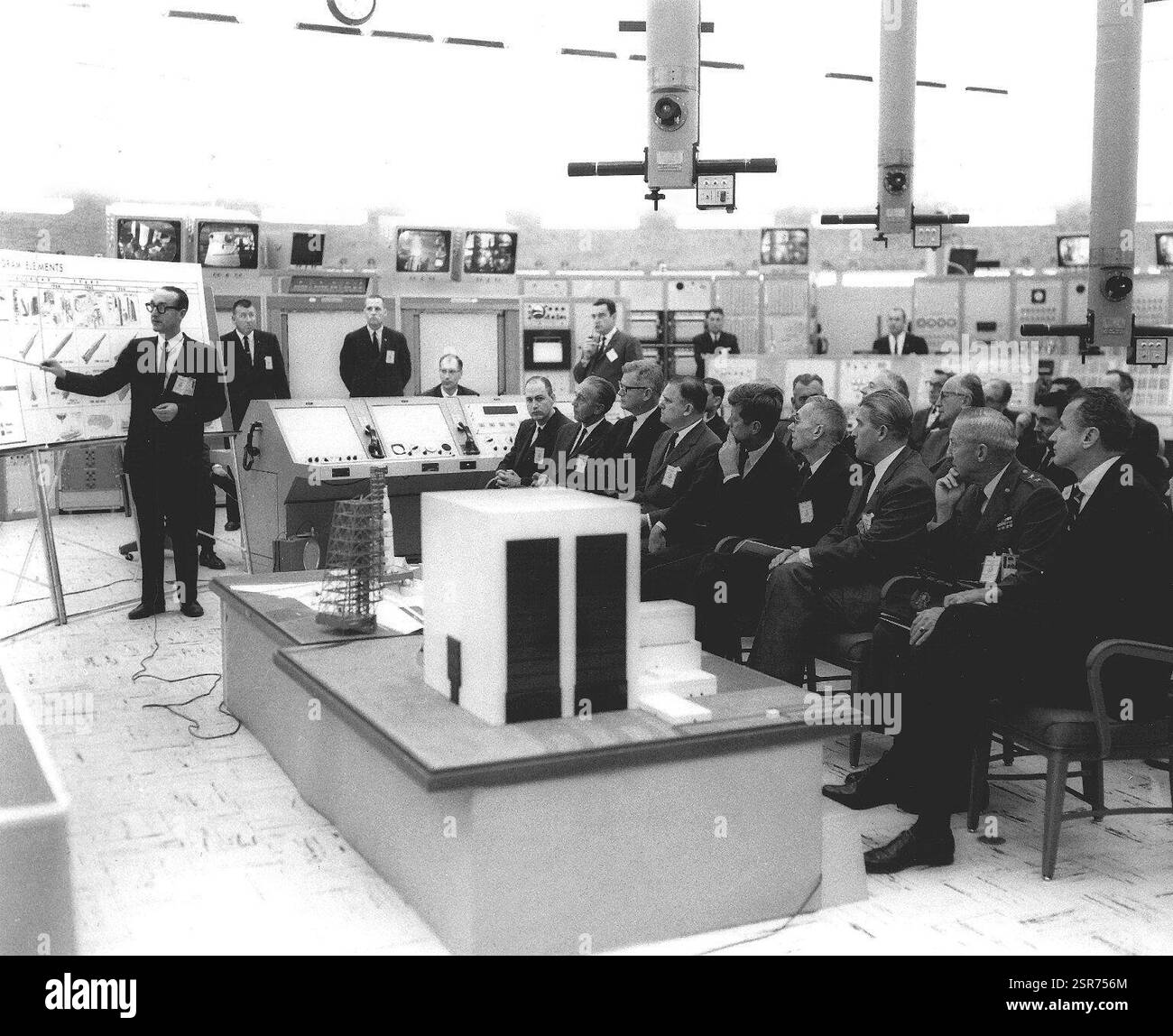 Präsident John F. Kennedy erhält ein Briefing über das Apollo-Programm, das ehrgeizige Projekt zur Landung eines Mannes auf dem Mond. Die ersten bemannten US-Weltraummissionen fanden während seiner Amtszeit statt, wobei Kennedy sich für die NASA einsetzte und das öffentliche Interesse an der Weltraumforschung förderte. Das Blockhaus, ein verstärkter, kuppelförmiger Bunker in der Nähe des Startplatzes, verwendete an der Decke montierte Periskope zur sicheren Raketenbeobachtung. Im Vordergrund zeigen Modellmodelle wichtige Apollo-Komponenten, darunter das Vertical Assembly Building, in dem Saturn-Raketen für ihre Mondmissionen zusammengebaut wurden. Stockfoto