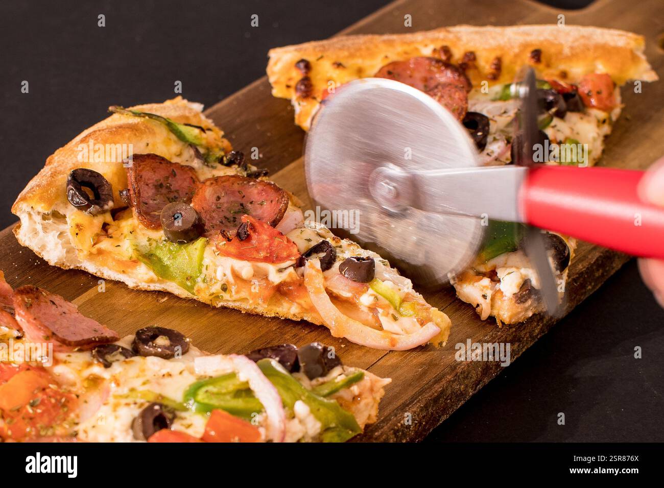 Schneiden Sie von Hand verschiedene Pizzastücke mit Pfefferoni, Oliven und Gemüse auf einem rustikalen Holzschneidebrett Stockfoto