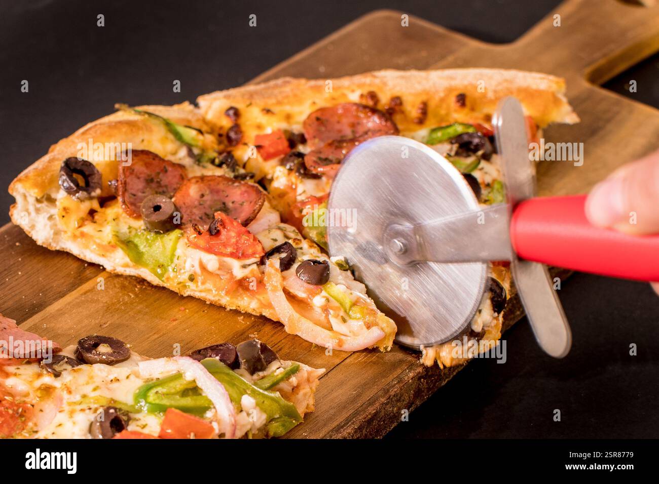 Schneiden Sie von Hand verschiedene Pizzastücke mit Pfefferoni, Oliven und Gemüse auf einem rustikalen Holzschneidebrett Stockfoto