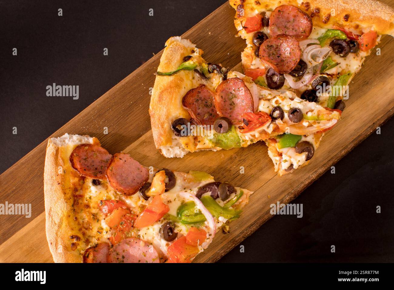 Scheiben von traditioneller Pizza mit Wurst, Pilzen, Paprika, Tomaten, Oliven, Käse und Zwiebeln auf einem rustikalen Holzschneidebrett Stockfoto