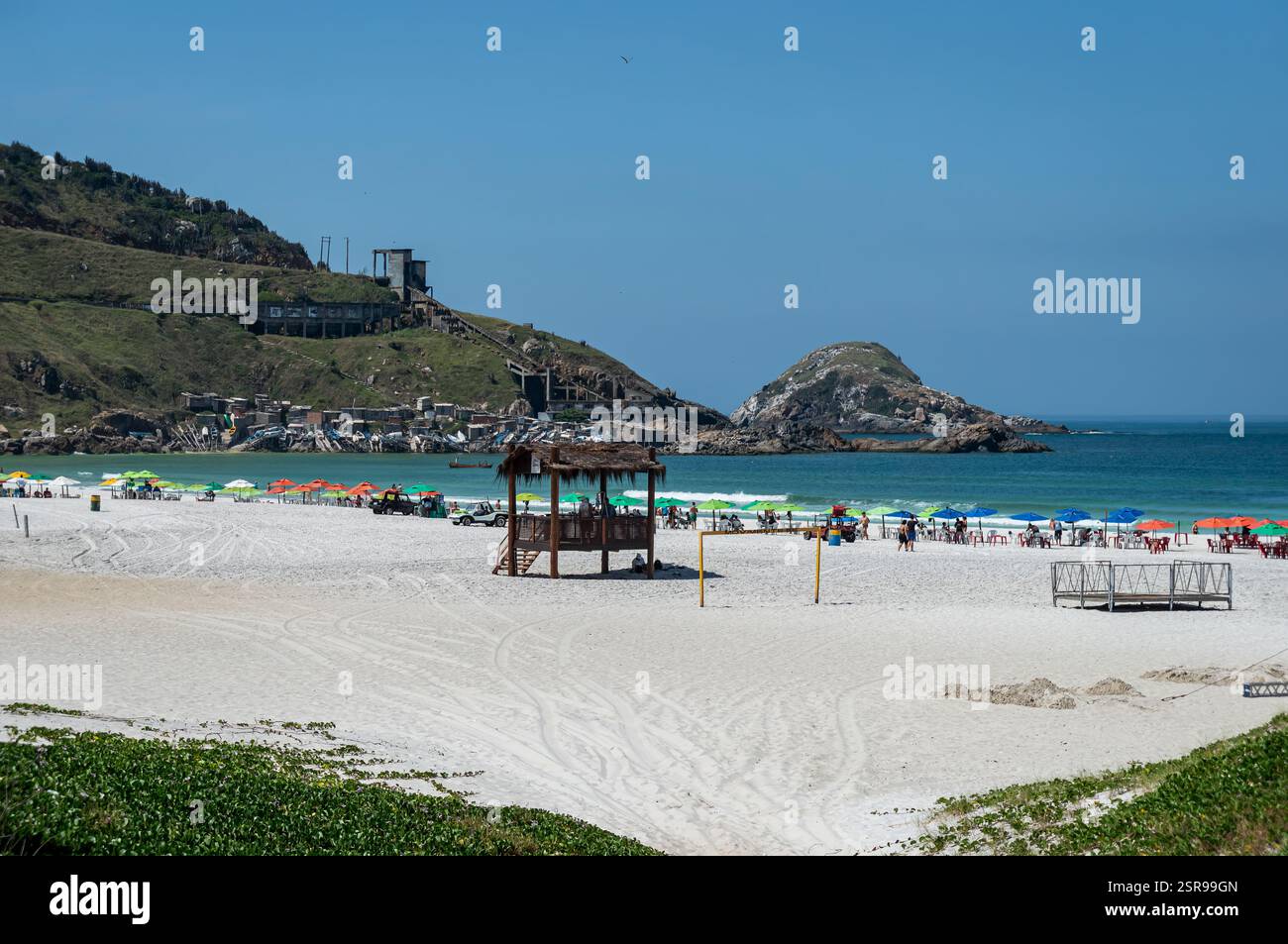 Südblick auf den Strand Praia Grande mit seinem weißen Sand, leuchtenden Sonnenschirmen und malerischen Hügeln unter einem hellen Sommermorgen sonnigen, klaren blauen Himmel. Stockfoto