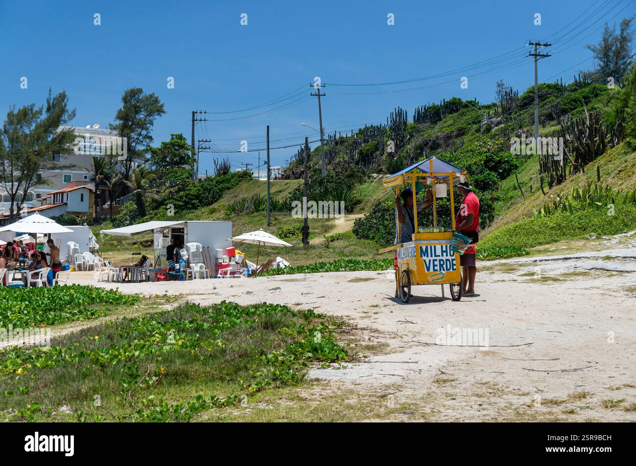Ein Straßenverkäufer, der unreife Mais am Strand von Prainha mit grünen Hügeln unter hellem und heißem Sommernachmittag verkauft. Stockfoto