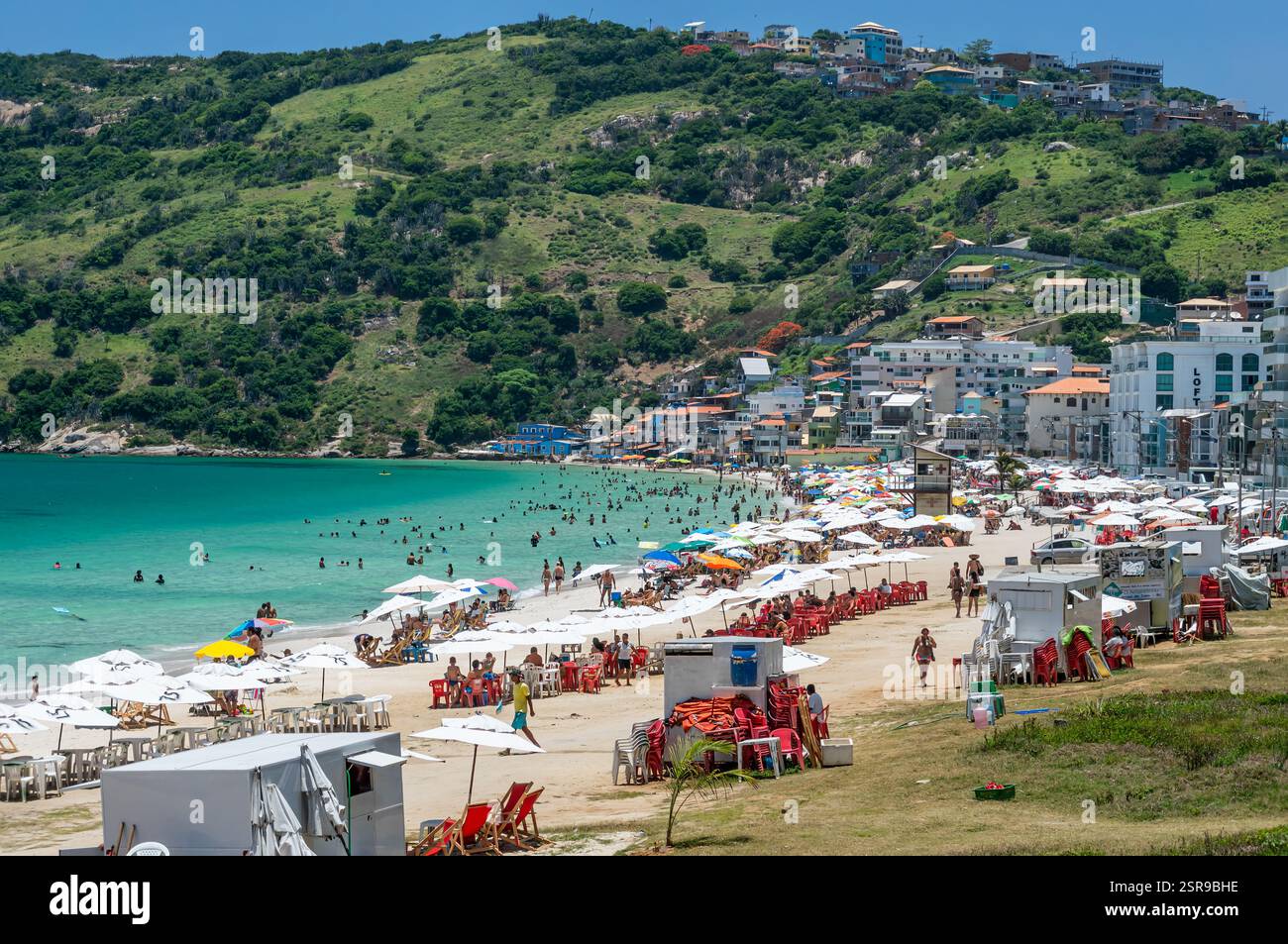 Die belebte Strandlinie von Prainha liegt südöstlich mit farbenfrohen Sonnenschirmen, Gebäuden und Küstenhügeln unter dem klaren blauen Himmel am Sommernachmittag. Stockfoto