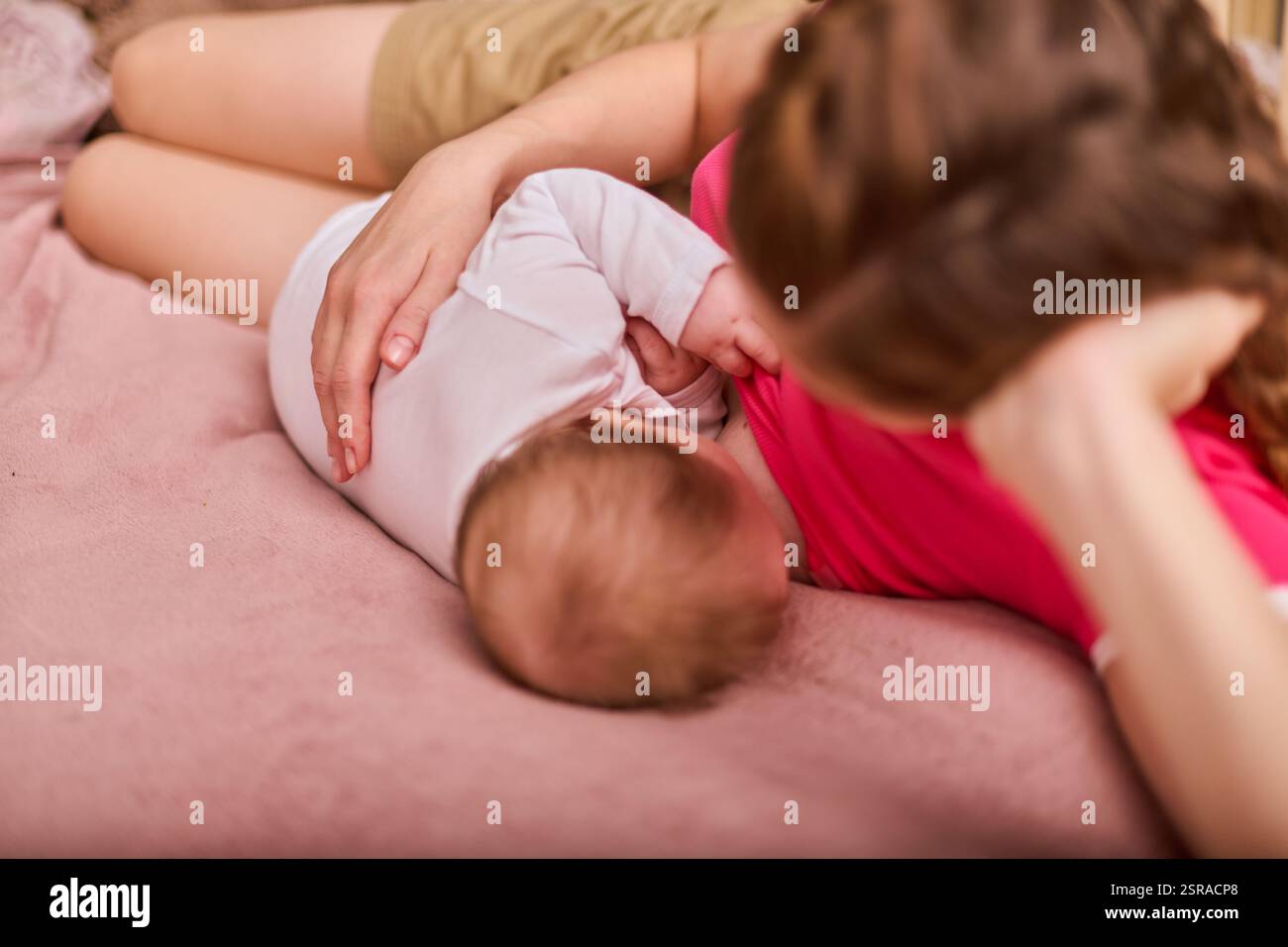 Junge Erwachsene mit geflochtenem Haar, gestilltes Kind auf rosa Tagesdecke. Intime und pflegende Atmosphäre mit sanfter Beleuchtung, die Bindungen einfängt Stockfoto