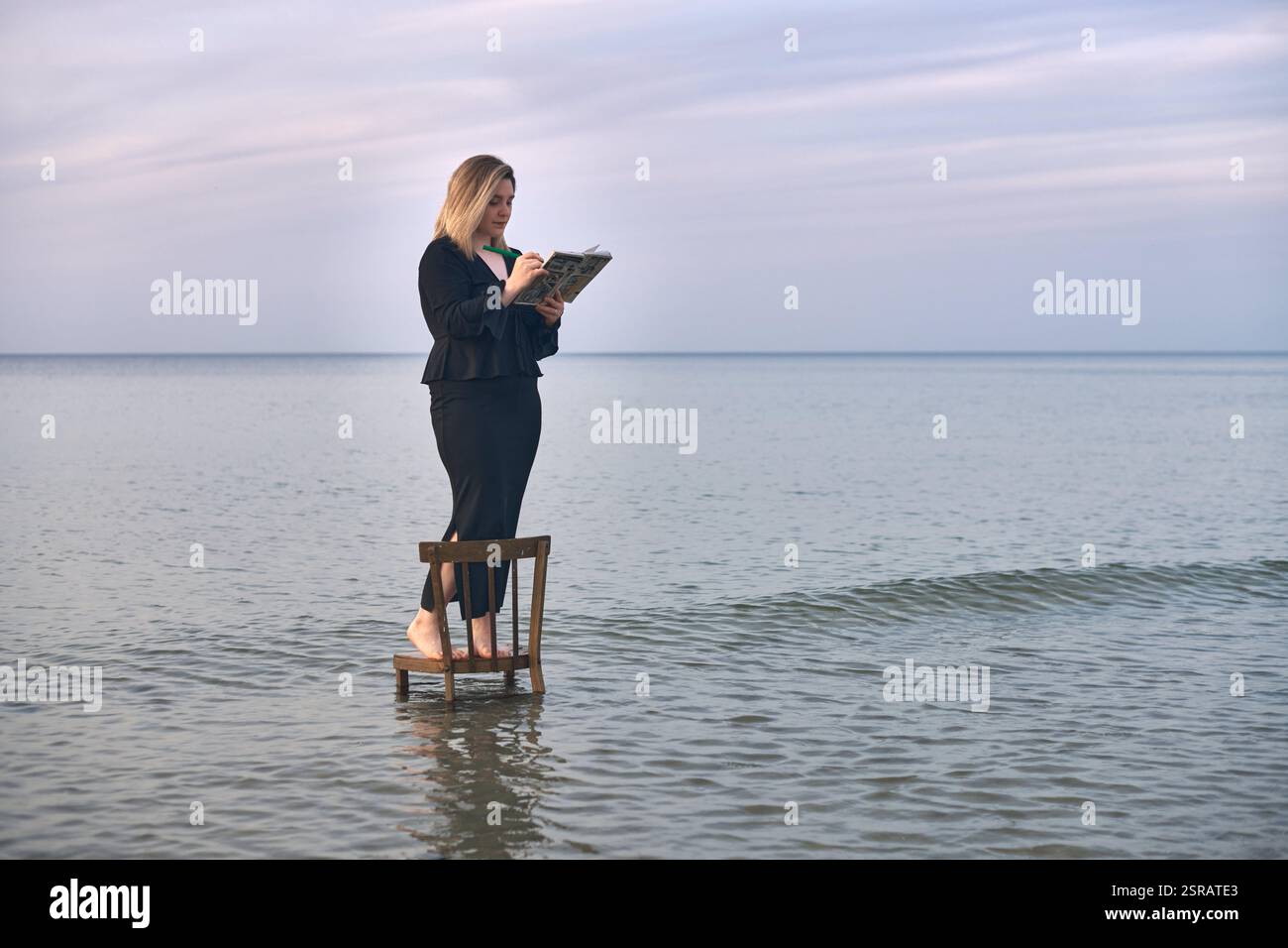 Junge Erwachsene Frau steht auf Holzstuhl in ruhigem Strandwasser und liest ein Buch. Ruhiger Himmel und sanfte Wellen schaffen eine friedliche und doch surreale Atmosphäre, Symbol Stockfoto
