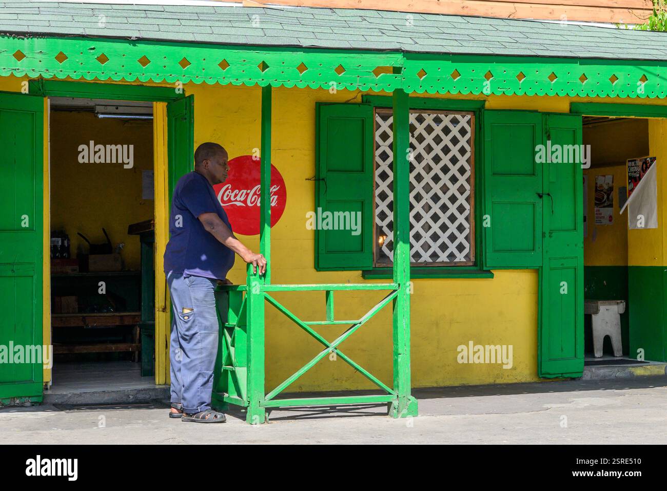 Cafe Worker steht vor seinem traditionellen kleinen lokalen Cafe mit schnörkelloser Basic Front, außer Einem Coca Cola Schild an der Bay Road Basseterre St Kitts Stockfoto