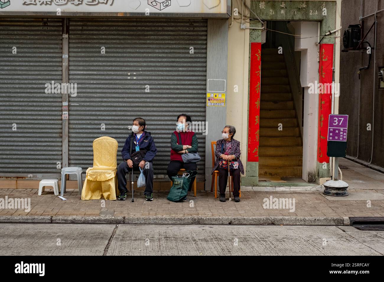 Ältere Frauen sitzen draußen auf Stühlen und warten auf den grünen Kleinbus in AP Lei Chau, der traditionelle und moderne Elemente in einer ruhigen urbanen Szene vereint. Stockfoto