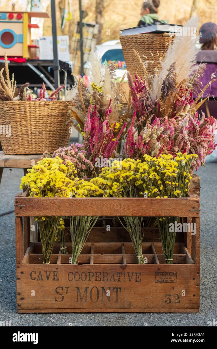 Eine rustikale Holzkiste voller lebendiger Blumen auf einem Markt im Freien, umgeben von Korbkörben und handgefertigten Arrangements. Montbrun-Bocage, Frankreich Stockfoto