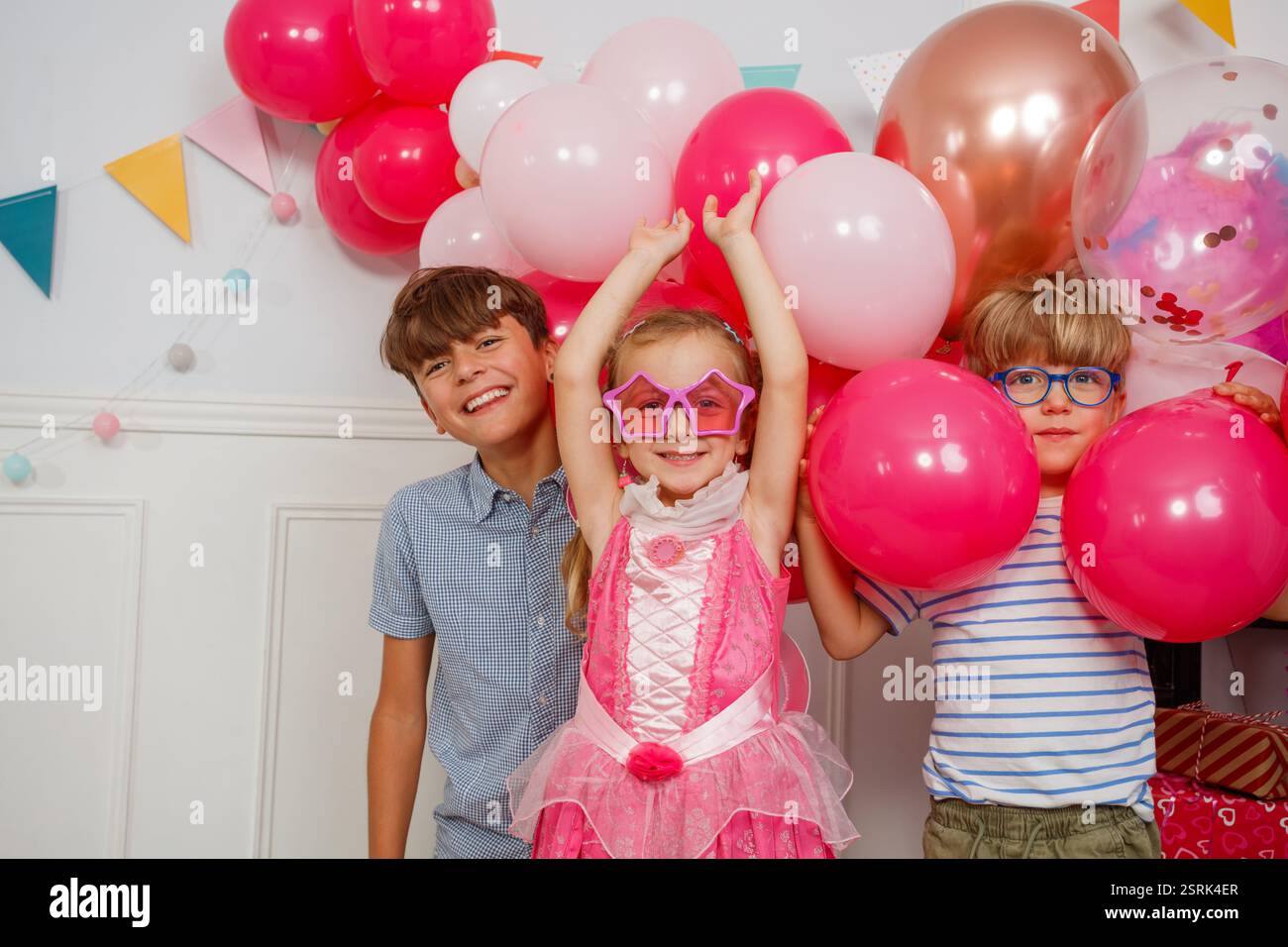 In einer fröhlichen Szene lachen drei Kinder neben hellen Ballons und verschiedenen Partybannern Stockfoto