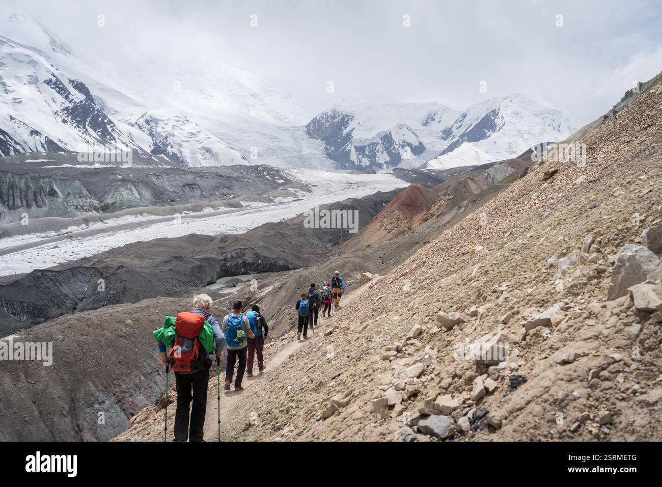 Touristen wandern durch felsiges Gelände mit Gletschern und schneebedeckten Gipfeln im Hintergrund, Pamir, Kirgisistan Stockfoto