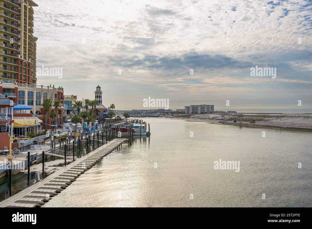 Das ruhige Wasser von Destin Harbor reflektiert die Wolken, mit einem Fußweg, einem Bootssteg und Ferienwohnungen am Ufer unter dem weiten Himmel. Stockfoto