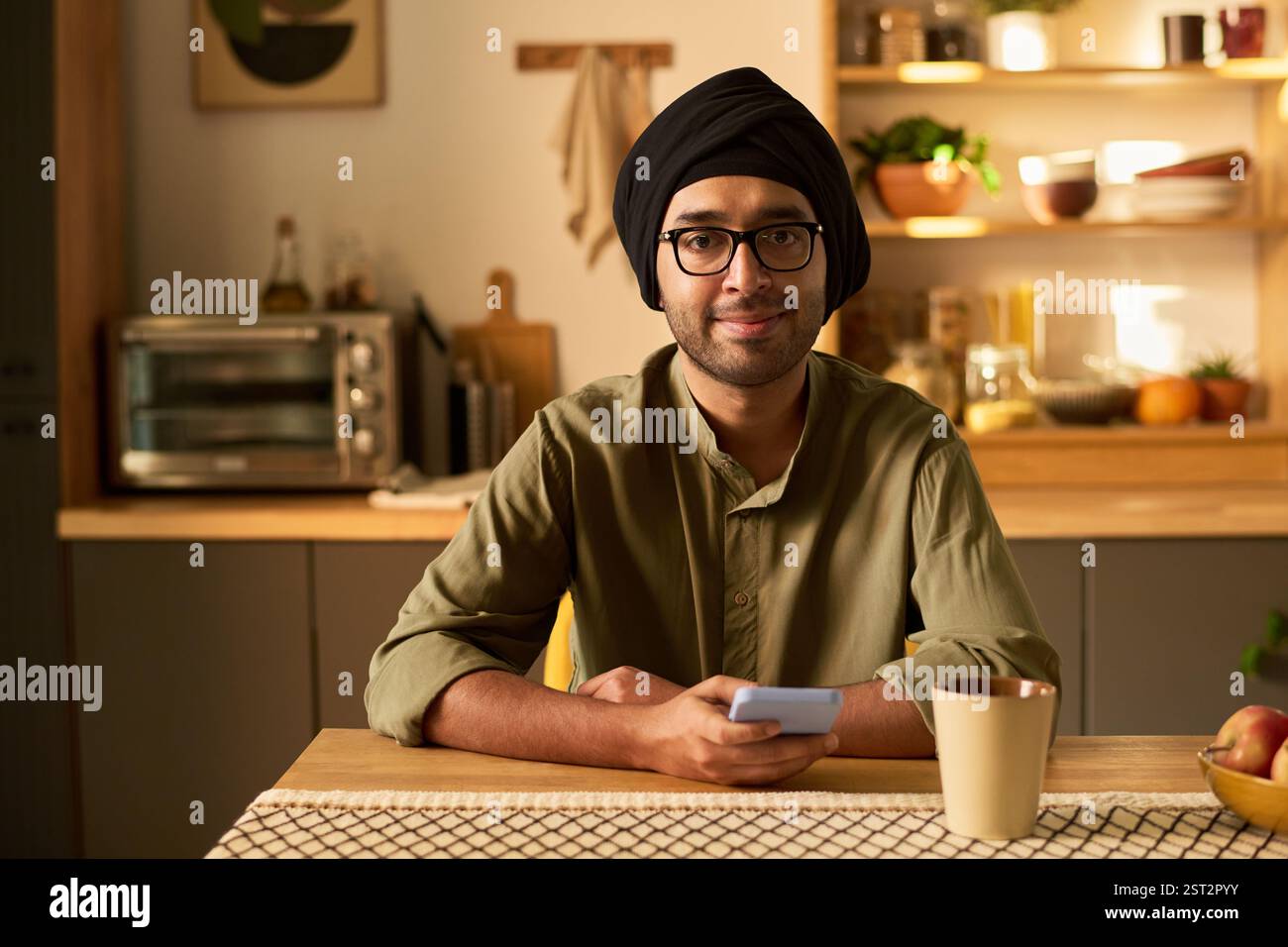 Porträt eines jungen Mannes mit einer Brille, der Turban trägt, am Küchentisch sitzt, mit einem Tablet in den Händen warm lächelt, direkt in die Kamera blickt und eine freundliche Atmosphäre schafft Stockfoto