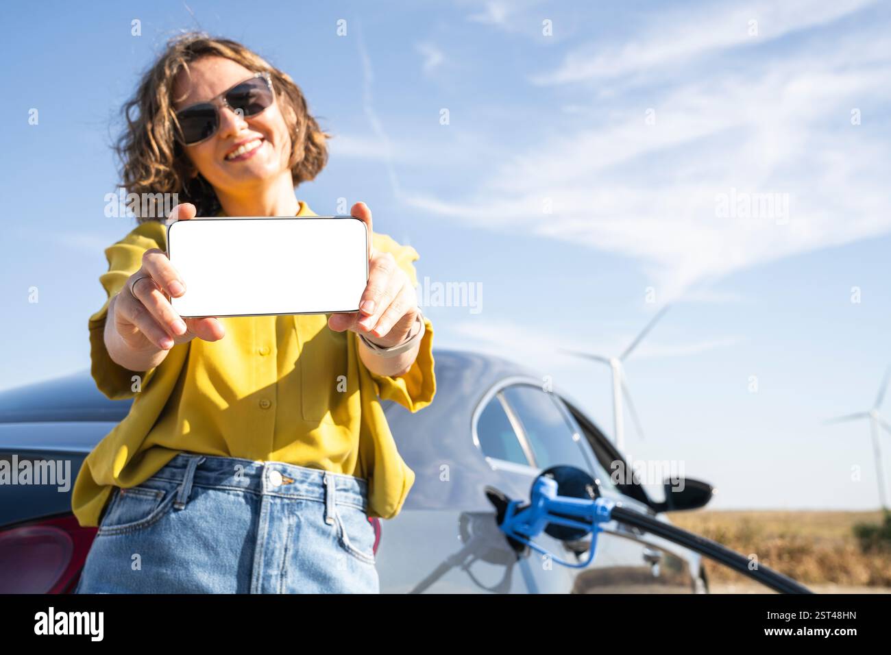 Frau mit Smartphone betankt Wasserstoffauto. Betanken des Autos mit Wasserstoffkraftstoff. Windräder im Hintergrund. Stockfoto