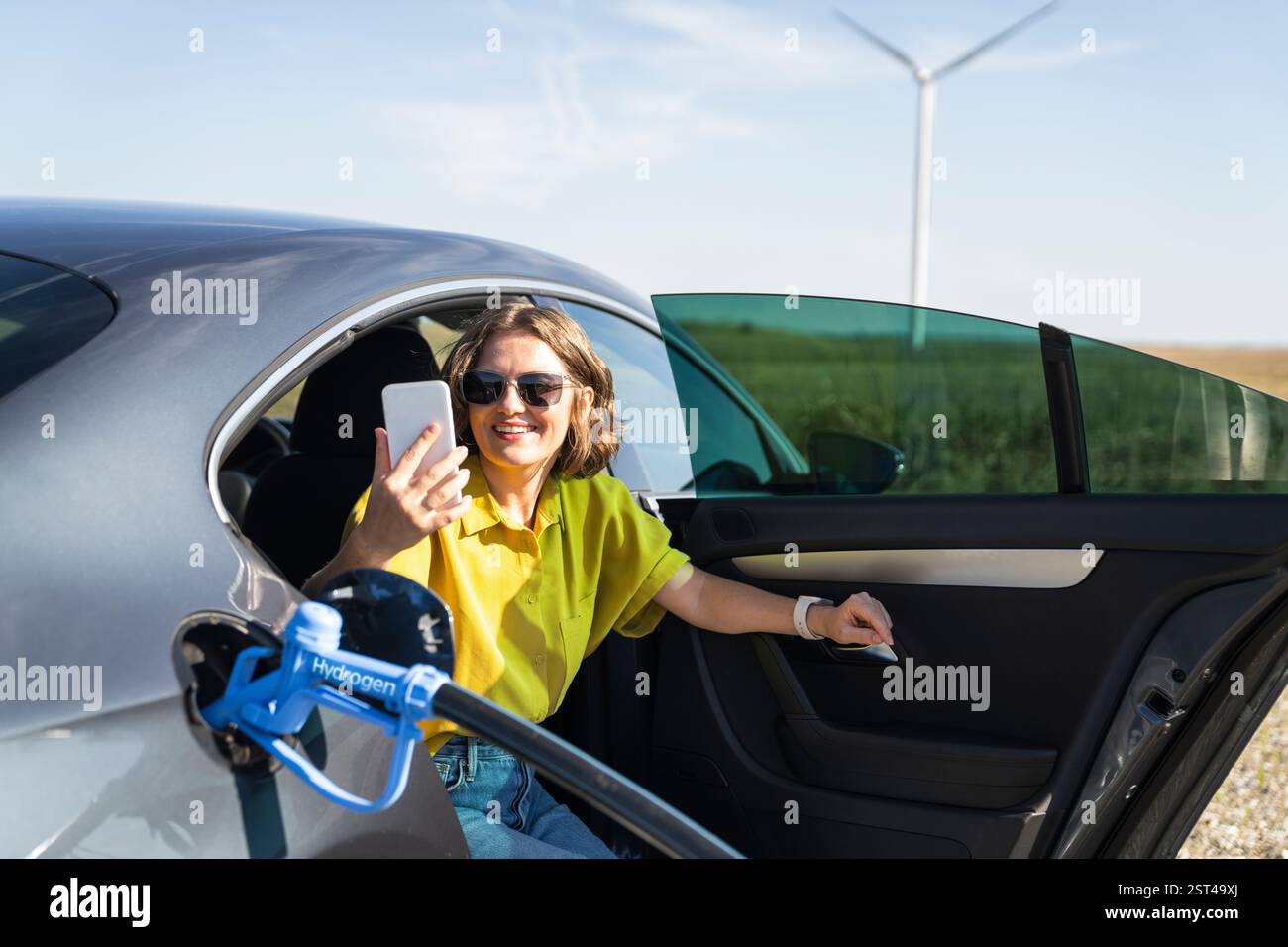 Frau mit Smartphone betankt Wasserstoffauto. Betanken des Autos mit Wasserstoffkraftstoff. Windräder im Hintergrund. Stockfoto
