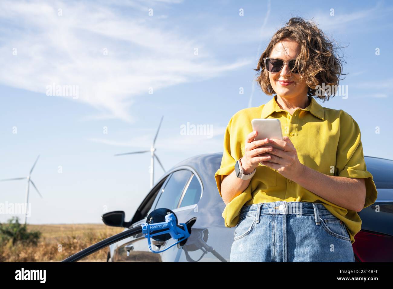 Frau mit Smartphone betankt Wasserstoffauto. Betanken des Autos mit Wasserstoffkraftstoff. Windräder im Hintergrund. Stockfoto