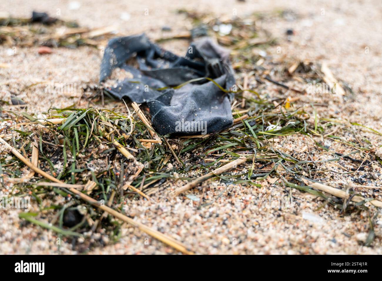 Ein Stück alten Stoffes, das auf dem Meer entsorgt wird, liegt am Sandstrand zwischen Algen und Muscheln, was die Abfälle der Textilindustrie veranschaulicht, die den Ozean verschmutzen Stockfoto