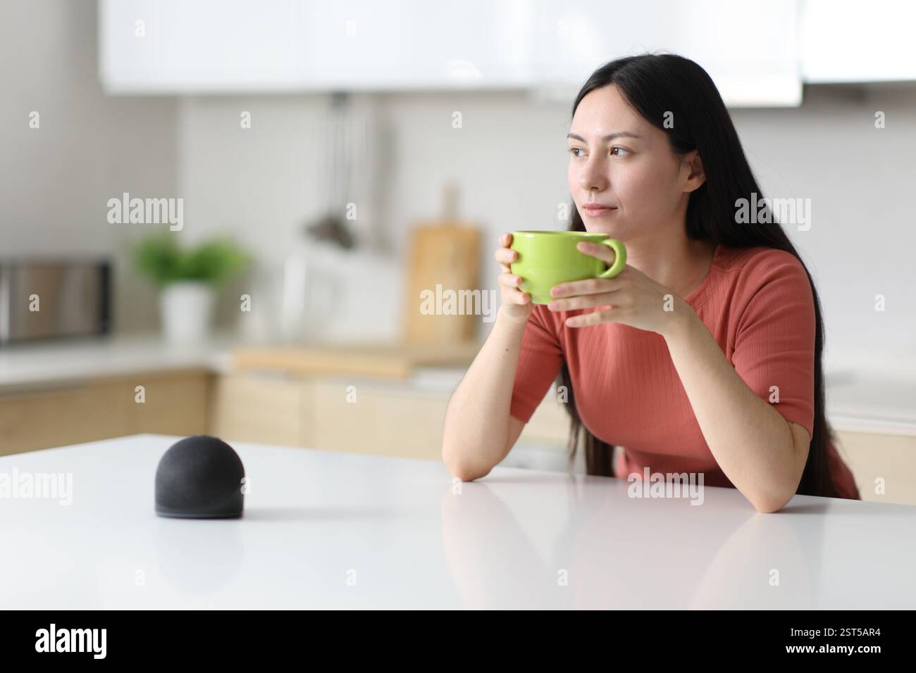 Entspannte asiatische Frau, die in einer Küche neben einem intelligenten Lautsprecher zu Hause trinkt Stockfoto